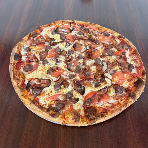 A whole Peoples Choice Pizza with bacon, sausage, and tomatoes on a wooden table.