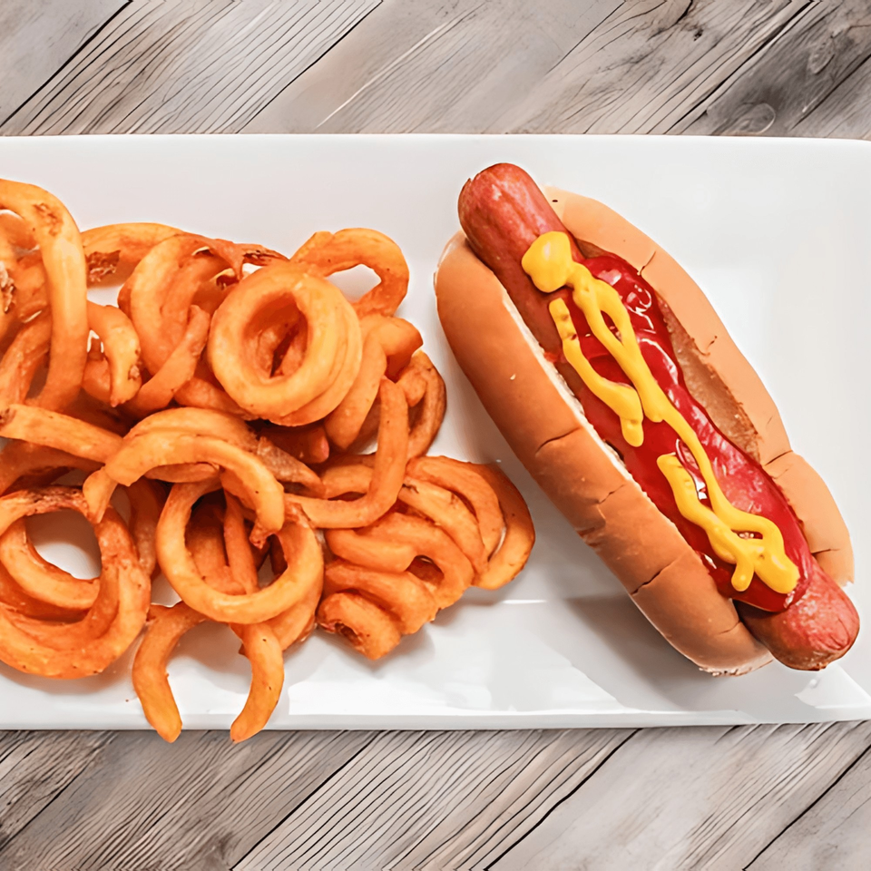 A hot dog with ketchup and mustard next to a pile of curly fries on a white plate.