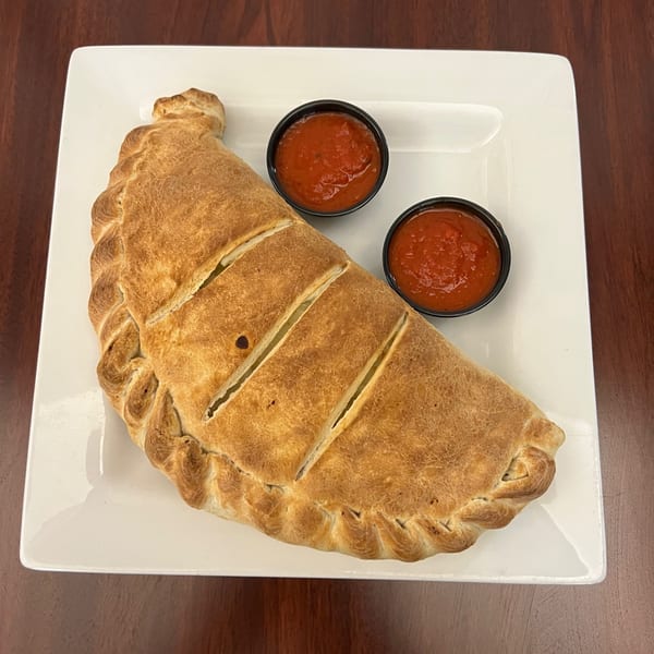A golden-brown calzone with three diagonal cuts, served with two small cups of marinara sauce on a white plate.