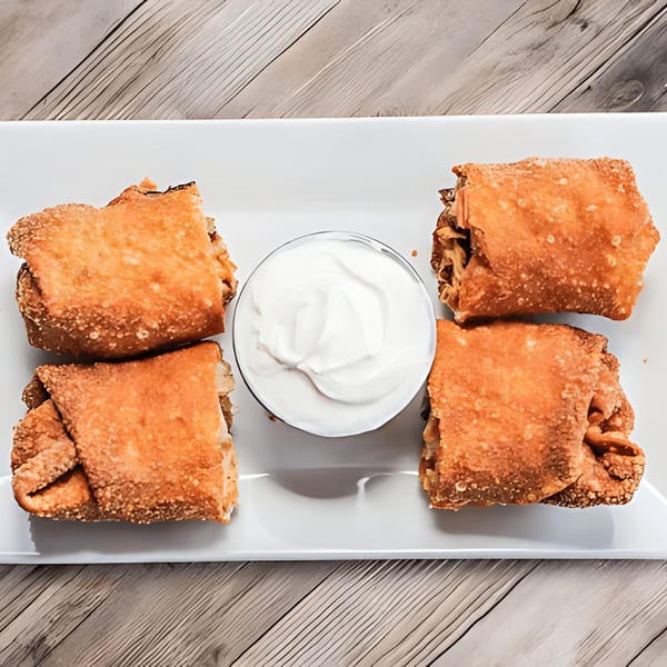 Four crispy egg rolls surround a small bowl of white dipping sauce on a white rectangular plate.